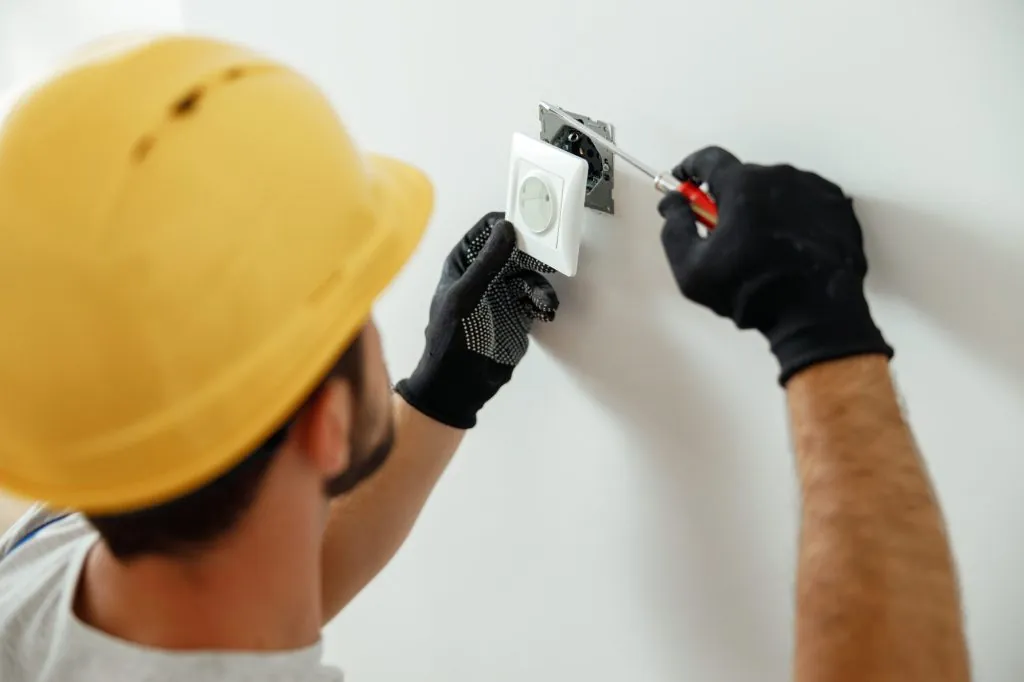 Eletricista de capacete amarelo e luvas pretas fixando uma tomada branca na parede com uma chave de fenda