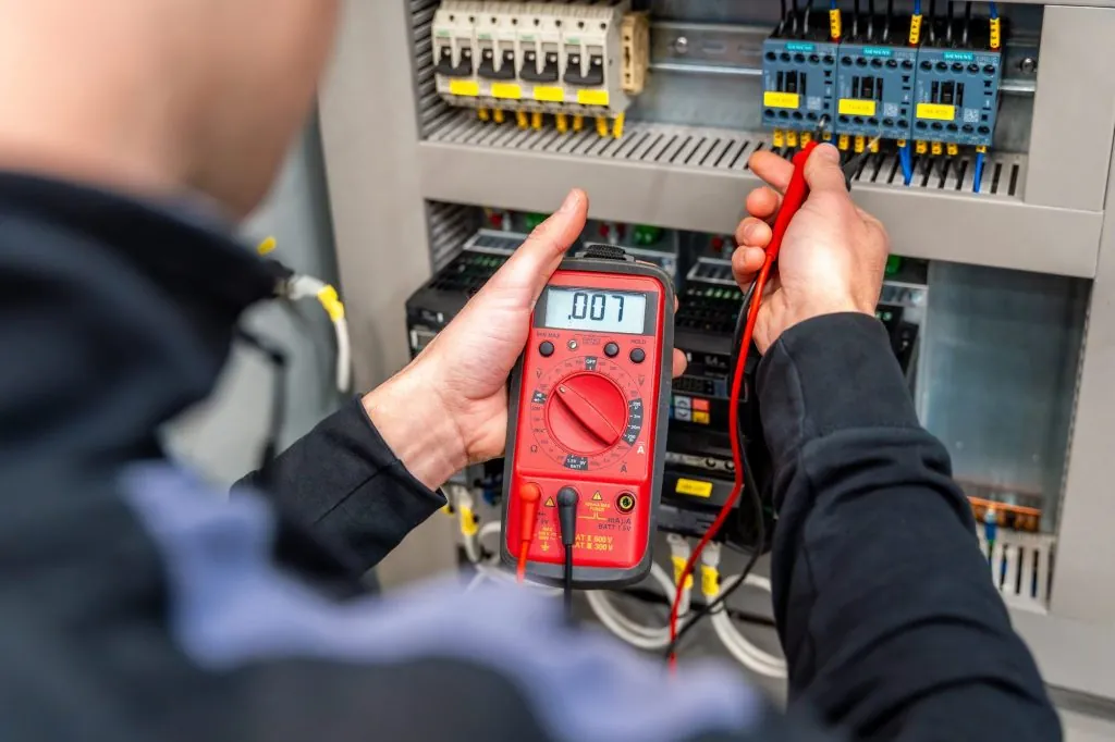 Eletricista medindo com um multímetro vermelho em um painel elétrico com disjuntores e contatores