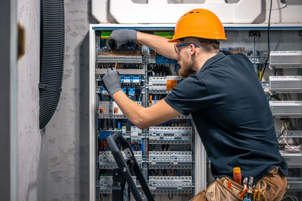 Eletricista de capacete laranja e luvas trabalhando em um grande quadro de distribuição, com disjuntores e fiação organizada